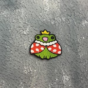 May include: Embroidered patch featuring a cartoon frog wearing a gold crown and a red and white spotted cape. The frog has a green body, a pink mouth, and black outlines. The patch is on a textured gray surface.