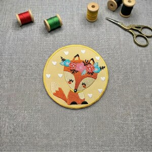 Peut inclure: Un écusson brodé circulaire représentant un renard orange stylisé avec une couronne de fleurs roses, rouges et bleues. Le renard est sur un fond jaune pâle décoré de petits cœurs blancs, entouré de fournitures de couture incluant du fil.