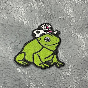 May include: A green frog wearing a black and white cow print hat with a pink star. The frog is sitting on a grey textured surface.