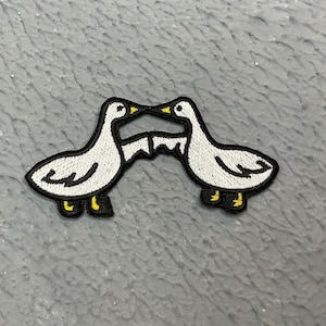 May include: Two white embroidered ducks facing each other with their beaks touching. The ducks have black outlines and yellow beaks.