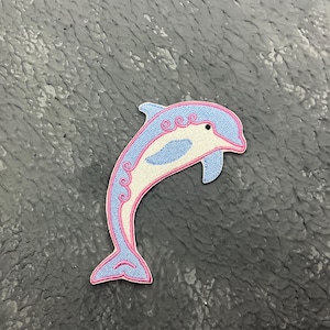 May include: A blue and white dolphin with pink outlines, embroidered on a gray fabric background.
