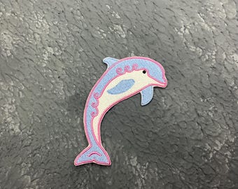 May include: A blue and white dolphin with pink outlines, embroidered on a gray fabric background.