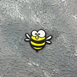 May include: A cartoon bee patch with a yellow and black striped body, white wings, and large white eyes. The patch is embroidered with black outlines and is set against a textured grey background.
