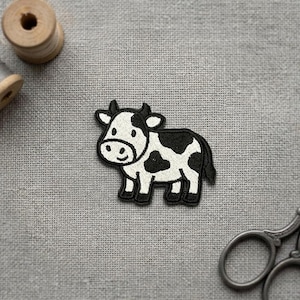 May include: A black and white cow patch with a smiling face. The embroidered patch features a black outline and is set against a gray fabric background. A spool of thread and scissors are also visible.
