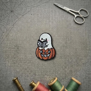 May include: An embroidered patch featuring a white ghost with pink cheeks reading a dark grey book while sitting inside a carved orange pumpkin. The patch is displayed alongside silver metal scissors and several spools of coloured sewing thread.