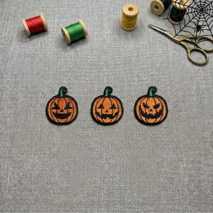 Peut inclure: Trois écussons brodés en forme de citrouille avec un tissu orange, des visages de jack-o'-lantern cousus en noir et des tiges vertes. Les écussons sont disposés horizontalement avec des bobines de fil rouge, vert, jaune et des ciseaux en métal.