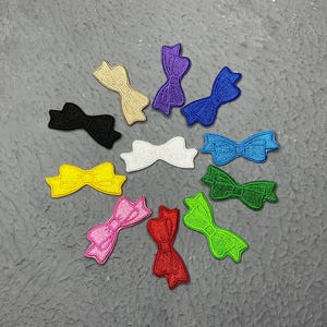 May include: Assortment of colourful felt bow appliques in various colours, including pink, blue, yellow, green, white, and black. The bows are all in the shape of a classic bow with a centre knot.