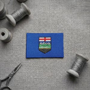 Peut inclure: Un écusson rectangulaire brodé représentant le drapeau bleu de l'Alberta avec un écusson central affichant une croix rouge, des montagnes blanches, des collines vertes et un champ de blé doré, entouré de bobines de fil et de ciseaux.