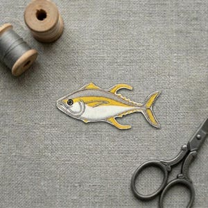 May include: An embroidered fish patch featuring a yellow and white body with grey detailing. The patch is positioned near a wooden spool of grey thread and a pair of metal scissors, showcasing a detailed design of a tuna-like fish with fins.