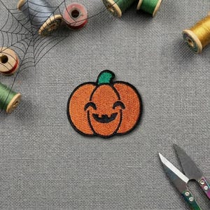 Peut inclure: Un écusson brodé en forme de citrouille avec un corps orange vif, une tige vert foncé et un visage de jack-o'-lantern cousu en noir. L'écusson est entouré de diverses bobines de fil à coudre et d'une paire de ciseaux en métal sur un tissu gris.