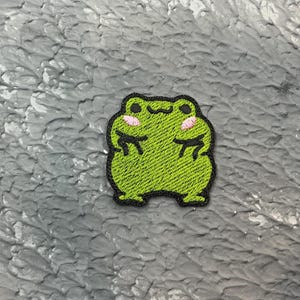 May include: A green embroidered frog patch with a black border. The frog has black arms and legs, and pink cheeks. The patch is on a textured grey surface.