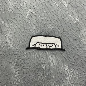 May include: A white embroidered patch with two cartoon faces peeking out from behind a white rectangle. The patch has a black outline.