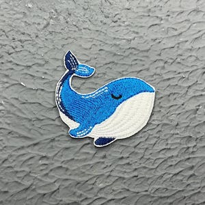 May include: A blue and white whale applique with a closed eye. The whale is embroidered with blue thread on a white fabric background.