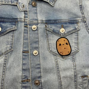 Op de afbeelding: Lichtblauwe spijkerjas met zilveren knopen en twee borstzakken. Een bruine, aardappelvormige patch met een lachend gezicht is op de linkerzak genaaid. De jas is gemaakt van een licht gewassen denim materiaal.