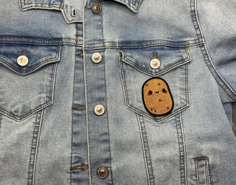 Op de afbeelding: Lichtblauwe spijkerjas met zilveren knopen en twee borstzakken. Een bruine, aardappelvormige patch met een lachend gezicht is op de linkerzak genaaid. De jas is gemaakt van een licht gewassen denim materiaal.