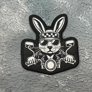 May include: A black and white patch depicting a rabbit on a motorbike. The rabbit is wearing sunglasses, a bandana, and a leather jacket. The patch shows the motorcycle's handlebars and front wheel in detail.