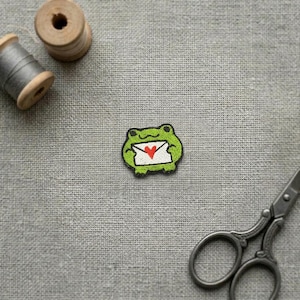 May include: An embroidered patch featuring a green frog holding a white envelope with a red heart on the front. The patch is positioned between a wooden spool of thread, a spool of grey thread, and a pair of dark metal sewing scissors.