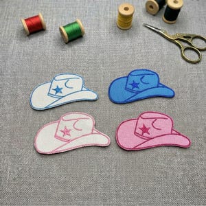 May include: Four embroidered cowboy hat patches in white, blue, and pink, each with a star and crescent moon design. The hats are surrounded by spools of thread and a pair of gold scissors.