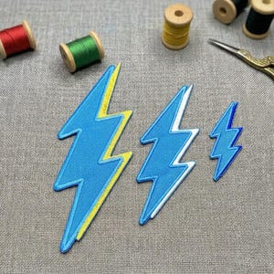 May include: Three blue lightning bolt patches with yellow and white trim, arranged on a gray fabric surface. Spools of thread in red, green, and yellow are visible, along with a pair of gold scissors. These patches are ideal for adding a unique touch to clothing or accessories.