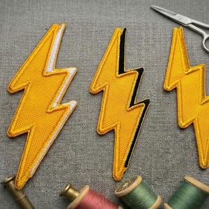 May include: Three yellow lightning bolt patches with white or black borders are arranged on a gray fabric surface. A pair of small silver scissors and spools of thread in various colors are also present, suggesting a sewing or crafting theme.