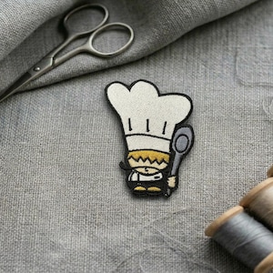 May include: Embroidered patch of a cartoon chef holding a spoon, wearing a white chef's hat and uniform. The patch is on a grey fabric background with sewing supplies, including scissors and spools of thread. The patch is approximately 5 cm tall.