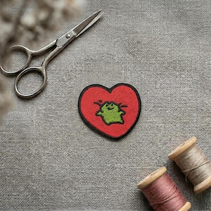 May include: A heart-shaped embroidered patch featuring a bright red background with a small, green, cartoon-style frog character in the centre. The patch has a thick black border and is positioned near a pair of silver metal scissors and two spools of thread.