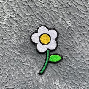 May include: A white flower with a yellow centre and a green stem and leaf. The flower is embroidered on a fabric patch.