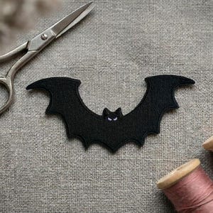 May include: A black embroidered bat patch with small white eyes, positioned next to a pair of silver metal sewing scissors and two wooden spools of thread, one with dusty pink thread and the other with light beige thread on a textured fabric.