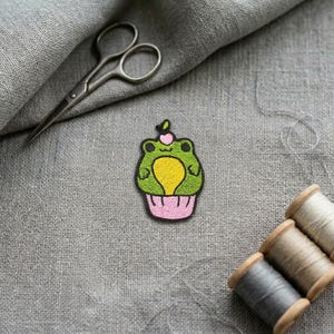 Peut inclure: Un écusson brodé représentant une grenouille verte en forme de cupcake avec un ventre jaune, des yeux noirs et une petite cerise rose sur le dessus. L'écusson est placé près d'une paire de ciseaux en métal et trois bobines de fil sur un tissu gris.