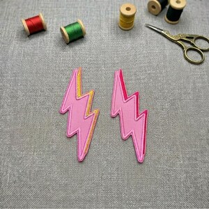 May include: Two pink lightning bolt patches with yellow and red detailing. The patches are embroidered and have a stitched outline. Spools of thread in red, green, yellow, and black are in the background, along with gold scissors.