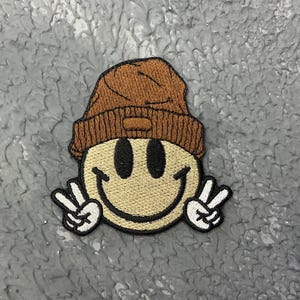 May include: Brown and beige embroidered smiley face patch with a brown beanie on top and white peace signs on each side.