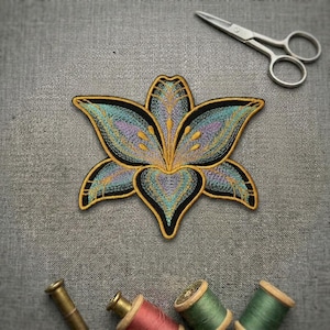 May include: An embroidered lily patch featuring teal, lavender, and gold thread with a black border. The patch is positioned below a pair of silver metal embroidery scissors and above three wooden spools of thread in red, green, and dark green.