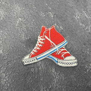 May include: A pair of red high-top sneakers patch with white laces, a white sole, and a blue stripe. The sneakers are embroidered with black stitching around the sole. The patch is on a gray surface.