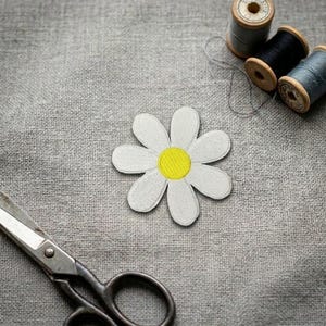 May include: A white daisy flower patch with a yellow center, positioned on a gray textured fabric surface. Nearby are three wooden spools of thread in black and gray tones, along with a pair of metal scissors with a silver finish.