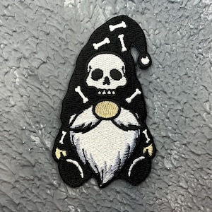 May include: Embroidered patch with a gnome design, featuring a skull face and crossed bones. The gnome wears a black hat with bone accents and has a white beard. The patch has a gold-coloured button on the chest and black boots.