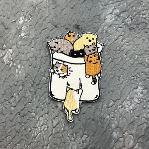 May include: Embroidered patch showing a white pocket filled with cartoon cats in shades of grey, orange, and yellow. One cat is hanging from the pocket. The patch is on a textured grey surface.