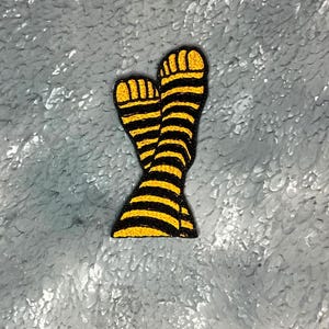 May include: Embroidered patch showing a pair of legs wearing black and yellow striped socks. The patch has a black border and is set against a textured grey background. The socks have yellow toes.