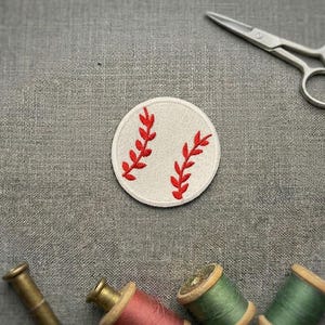 Peut inclure: Un écusson de baseball rond, crème, orné de coutures rouges. Il présente deux motifs rouges en forme de feuilles. Des ciseaux argentés et des bobines de fil de différentes couleurs sont également visibles.