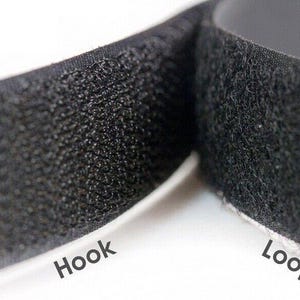 Op de afbeelding: Close-up van zwarte klittenbandstrips. De afbeelding toont de getextureerde oppervlakken van de haak- en luskanten, met de woorden "Hook" en "Loop" gedrukt op de witte achterkant.