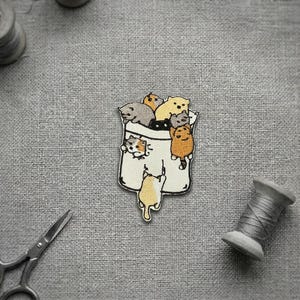 May include: Embroidered patch showing a white bag filled with cartoon cats in different colours. The cats are grey, orange, and yellow. Sewing tools, including scissors and thread spools, are around the patch on a grey fabric background.