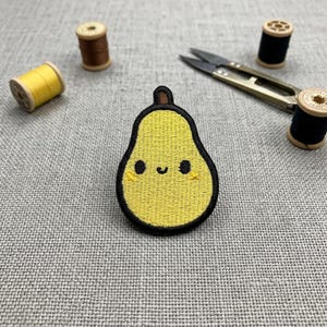 May include: A yellow pear-shaped embroidered patch with a smiling face and black outline. The patch is surrounded by spools of thread in yellow, brown, and black, along with small scissors, all on a grey fabric surface.