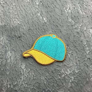 May include: Embroidered patch in the shape of a baseball cap. The cap has a turquoise crown and a yellow brim, outlined with gold thread. A small gold circle is at the top. The patch is likely for clothing or accessories.