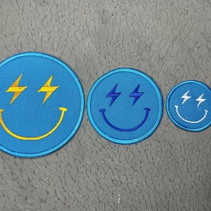 Peut inclure: Trois patchs brodés en forme de visage souriant avec des yeux en forme d'éclair. Le plus grand patch est bleu avec un sourire jaune et des éclairs jaunes. Le patch moyen est bleu avec un sourire bleu et des éclairs bleus. Le plus petit patch est blanc avec un sourire bleu et des éclairs bleus.