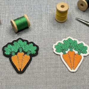 May include: Two carrot-shaped embroidered patches with green tops and orange carrots, alongside spools of red, green, and yellow thread, and gold scissors. The patches have black and white borders. The items are on a grey fabric surface.