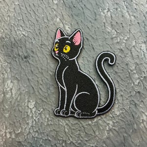 May include: Embroidered black cat patch with white outline, pink inner ears, and yellow eyes. The cat is sitting with its tail curled. The patch is on a textured grey surface.