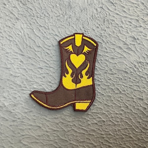 May include: A brown cowboy boot with a yellow heart and flame design. The boot is embroidered with yellow thread.