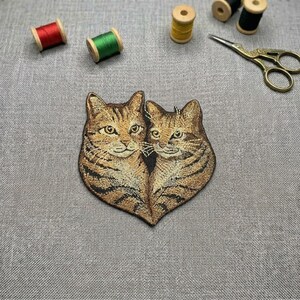 May include: Embroidered patch featuring two tabby cats, with brown and black stripes. The patch is heart-shaped. Sewing supplies, including spools of red, green, and yellow thread, and gold scissors are in the background.