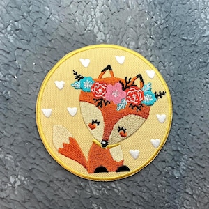 May include: Embroidered patch featuring a cartoon fox with a floral crown. The fox is orange and cream-coloured, with a yellow background and white heart accents. The patch has a yellow border.