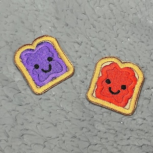 Peut inclure: Deux patchs brodés à repasser en forme de tranches de pain avec des visages souriants. Un patch a de la gelée violette et l'autre a de la gelée rouge.