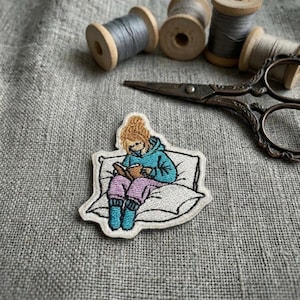 May include: An embroidered patch featuring a person with brown hair wearing a blue hoodie and purple trousers, sitting on a white pillow while reading a brown book. The patch is positioned near vintage metal scissors and several wooden thread spools.